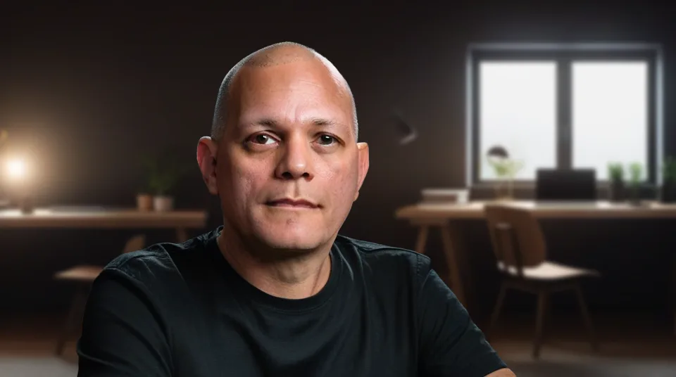 A generated presenter, a bald man in a black T-shirt, seated in a warmly lit home office with a window, a desk lamp and a chair behind him.