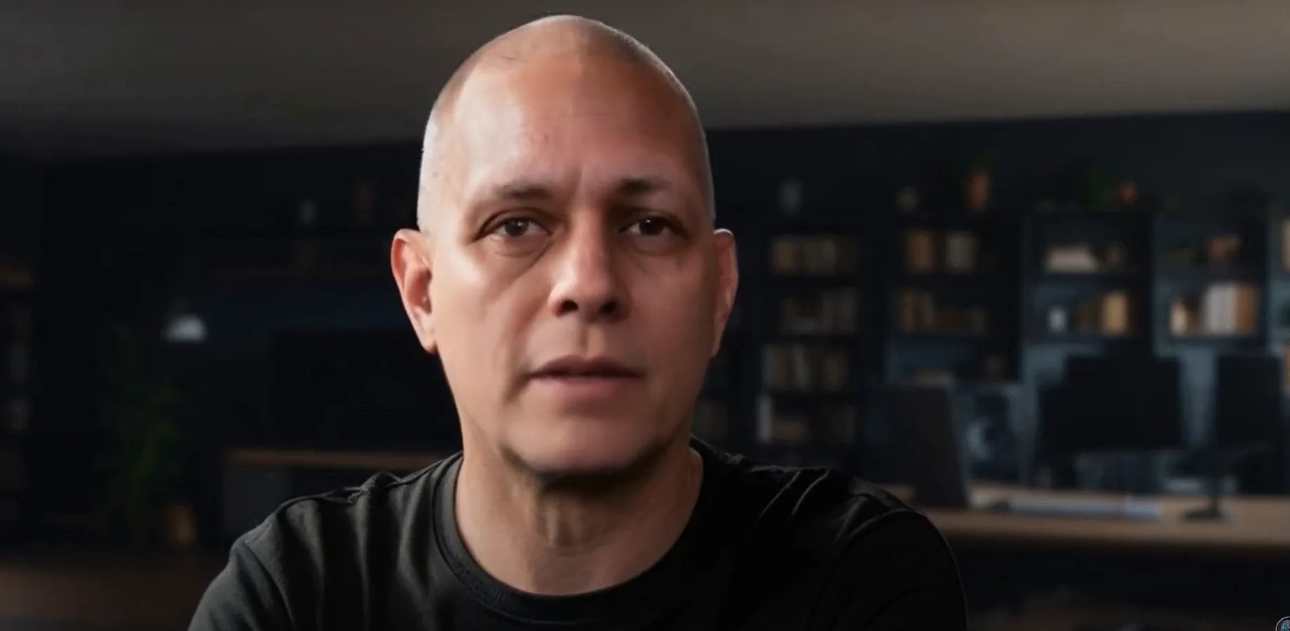 A lip-synced presenter avatar, a bald man in a black T-shirt, speaking to camera in front of dark bookshelves.