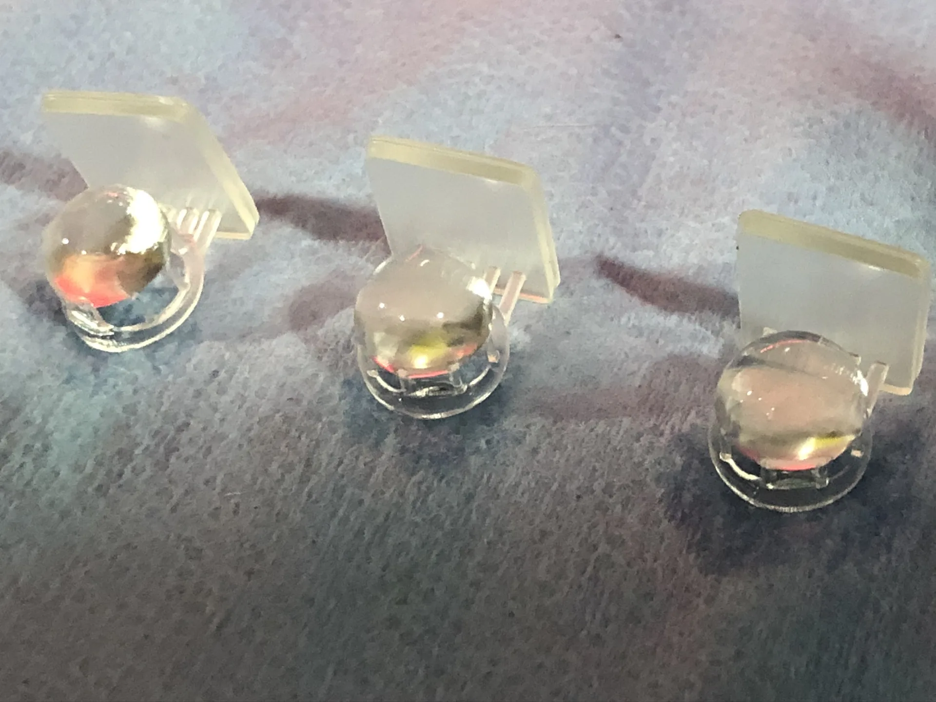Three small clear lenses with flat mounting tabs, printed in resin, sitting on a blue paper towel.
