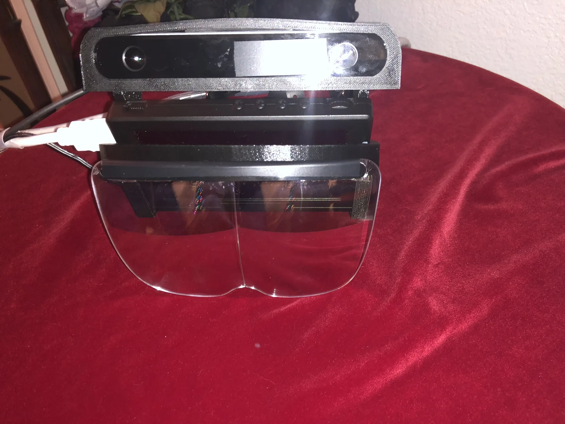 A black HoloFlect build resting on red velvet: a stereo camera on top, display housing below and a clear visor at the front.