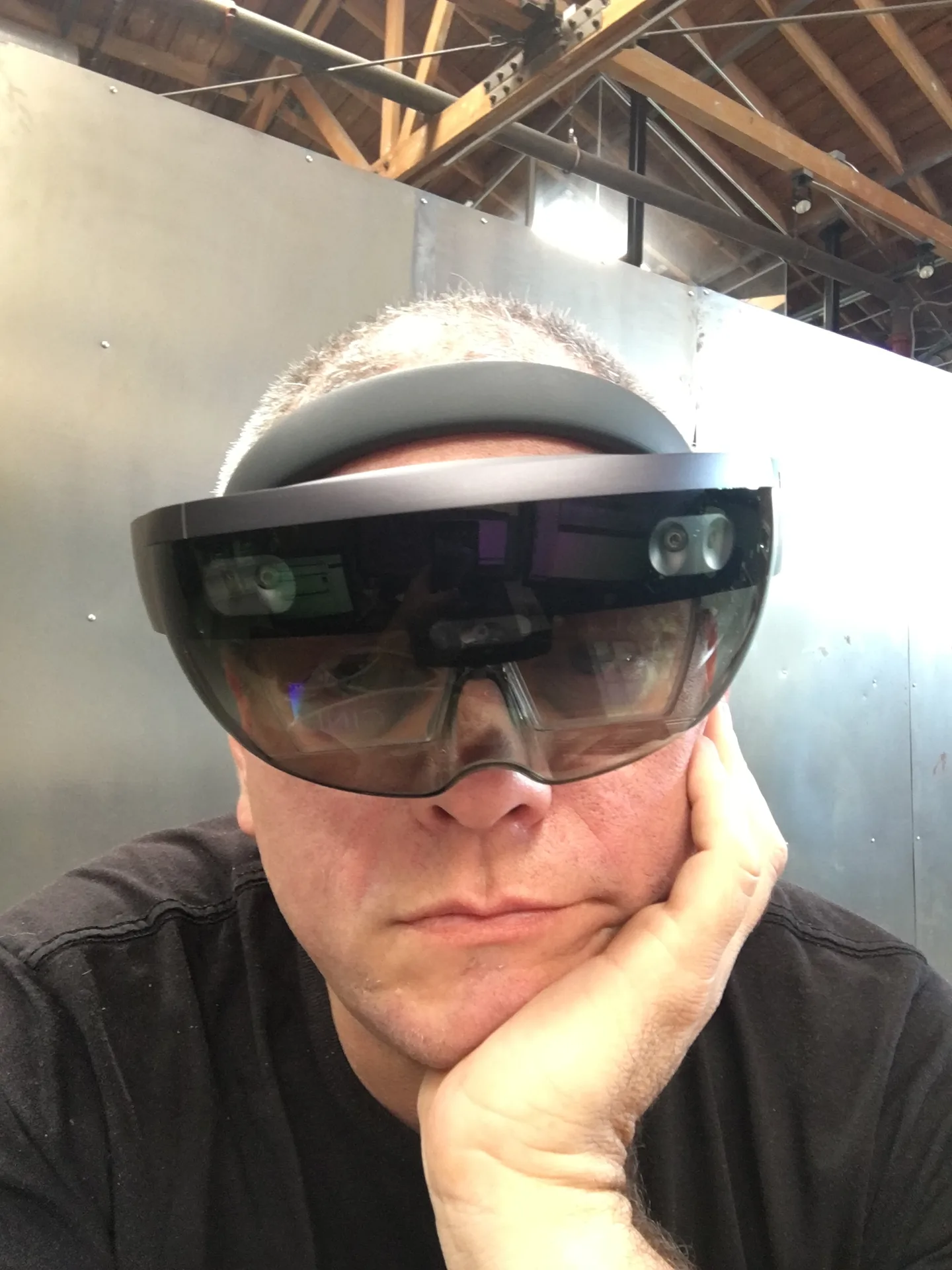 A man wearing a Microsoft HoloLens headset, chin resting on his hand, in front of a wall under exposed roof beams.