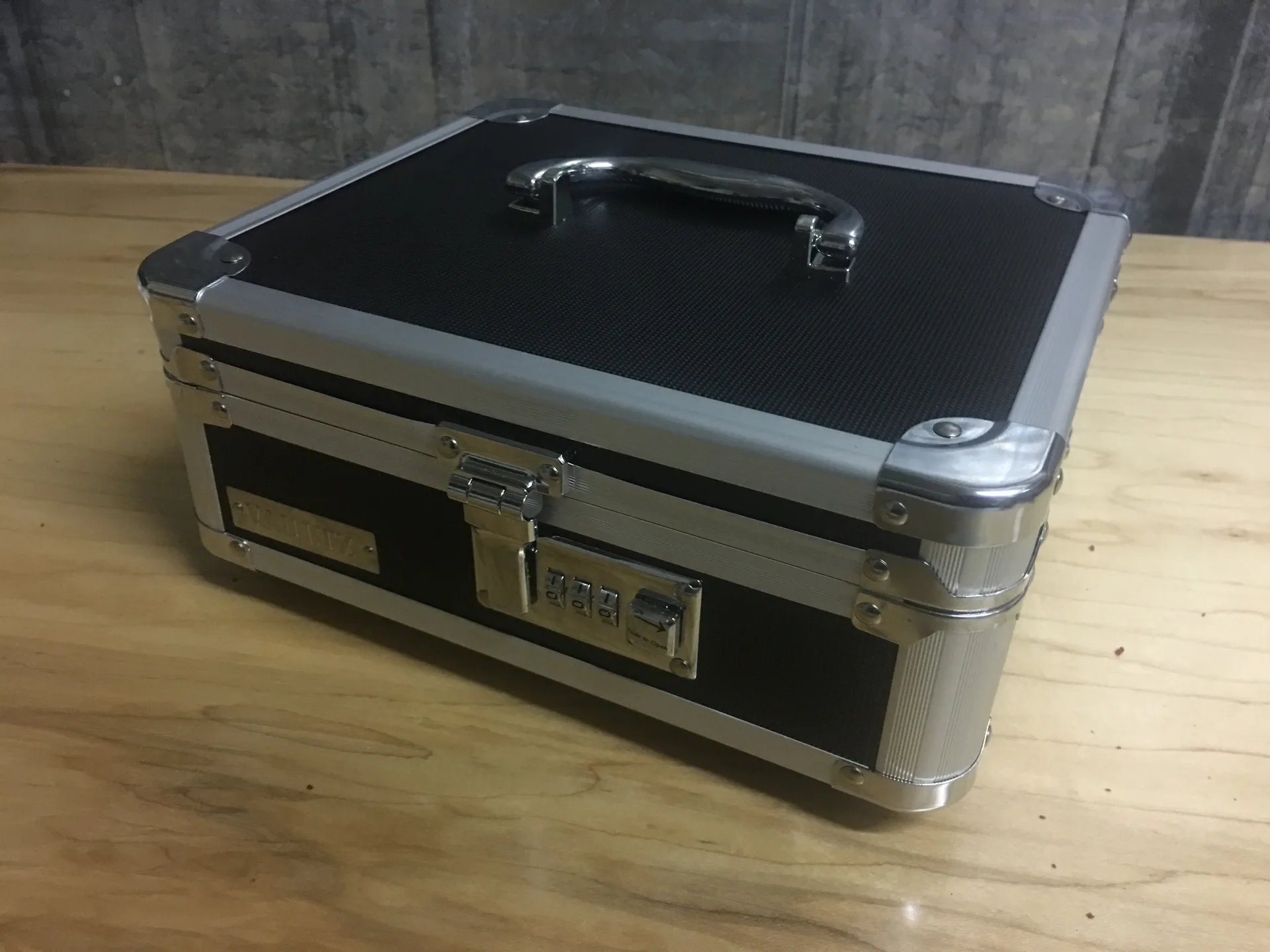 A closed black and aluminum carrying case with a handle and combination locks, on a wooden table.