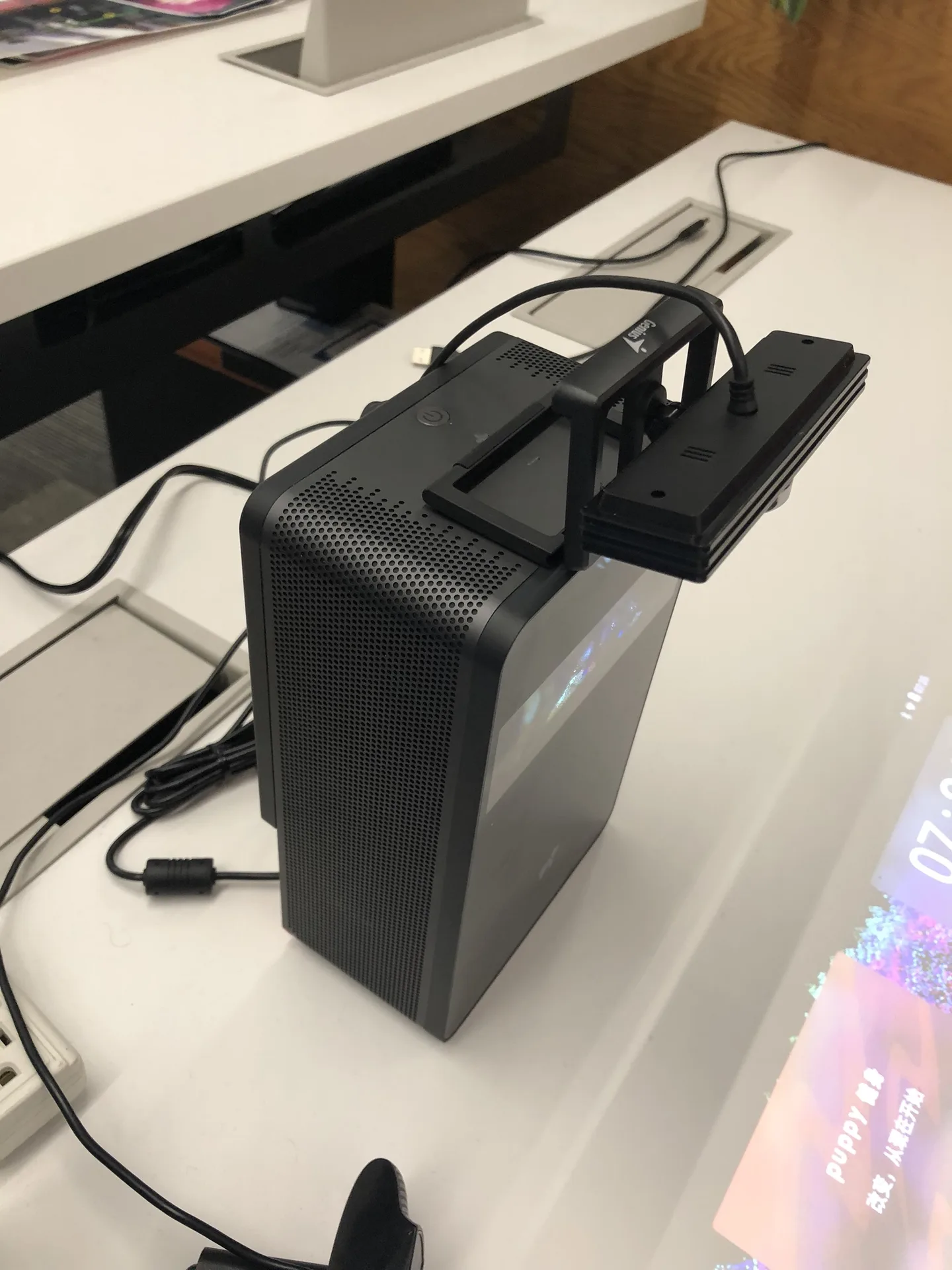 A compact black projector unit with a sensor bar mounted on its top, standing on a white desk with cables.
