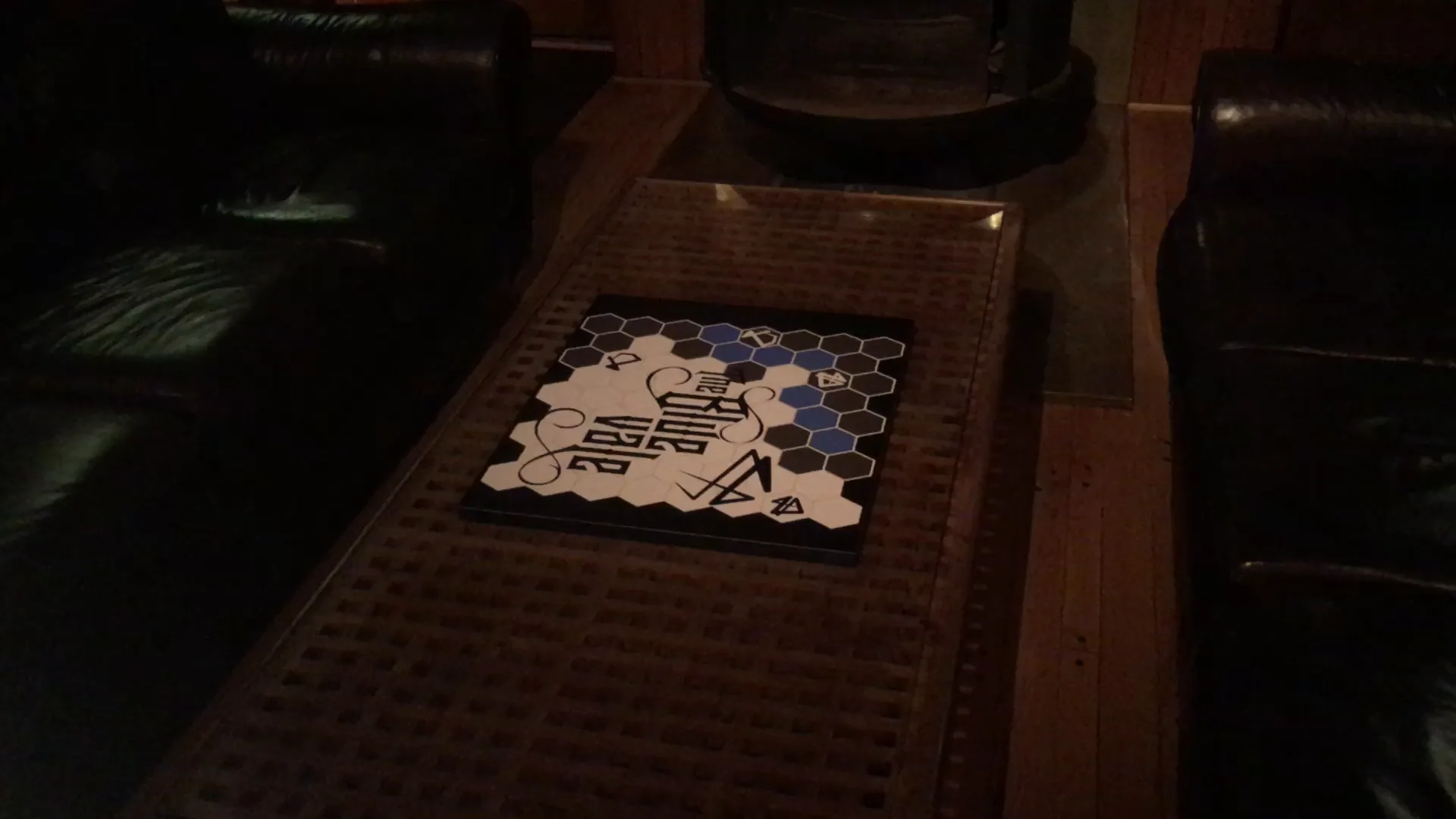 A small printed board with a hex pattern and The Blue Vale logo lying on a coffee table in a dim room.