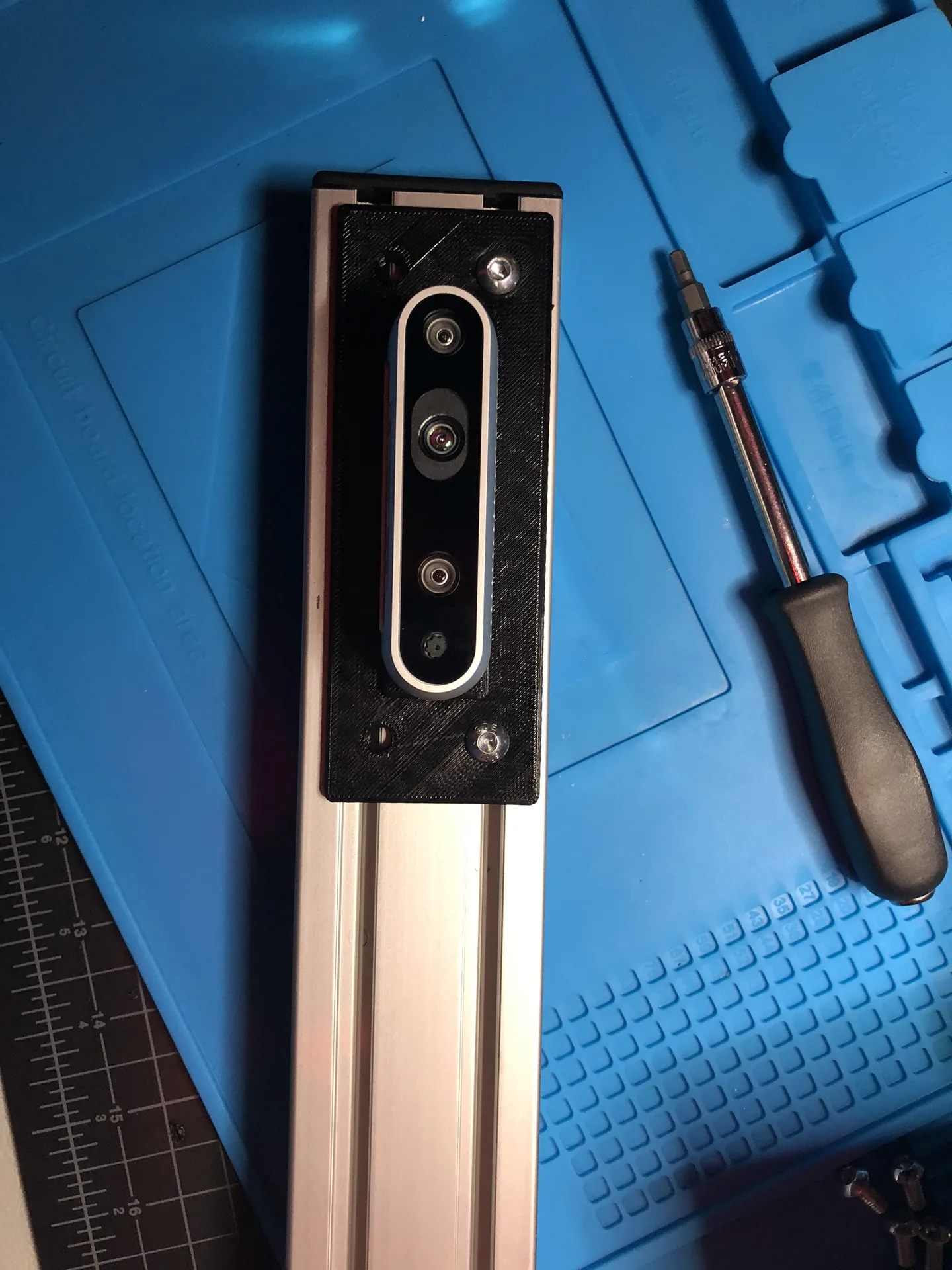 A RealSense camera fixed to a black printed plate at the end of an aluminum 80/20 rail, on a blue silicone mat.
